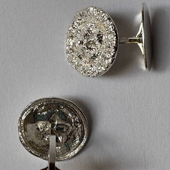 A pair of round organic cufflinks in silver, shown from both the front  and back (revealing a textured silver backing) on a neutral gray background.