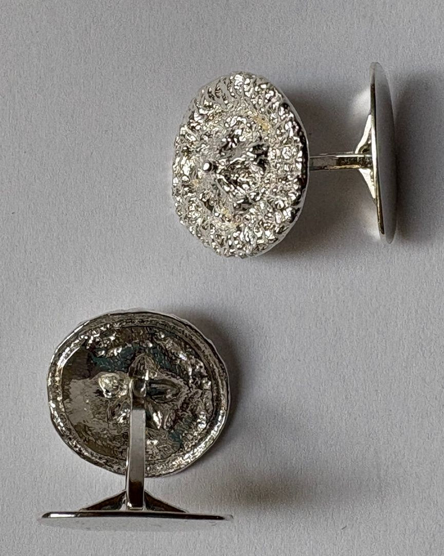 A pair of round organic cufflinks in silver, shown from both the front  and back (revealing a textured silver backing) on a neutral gray background.