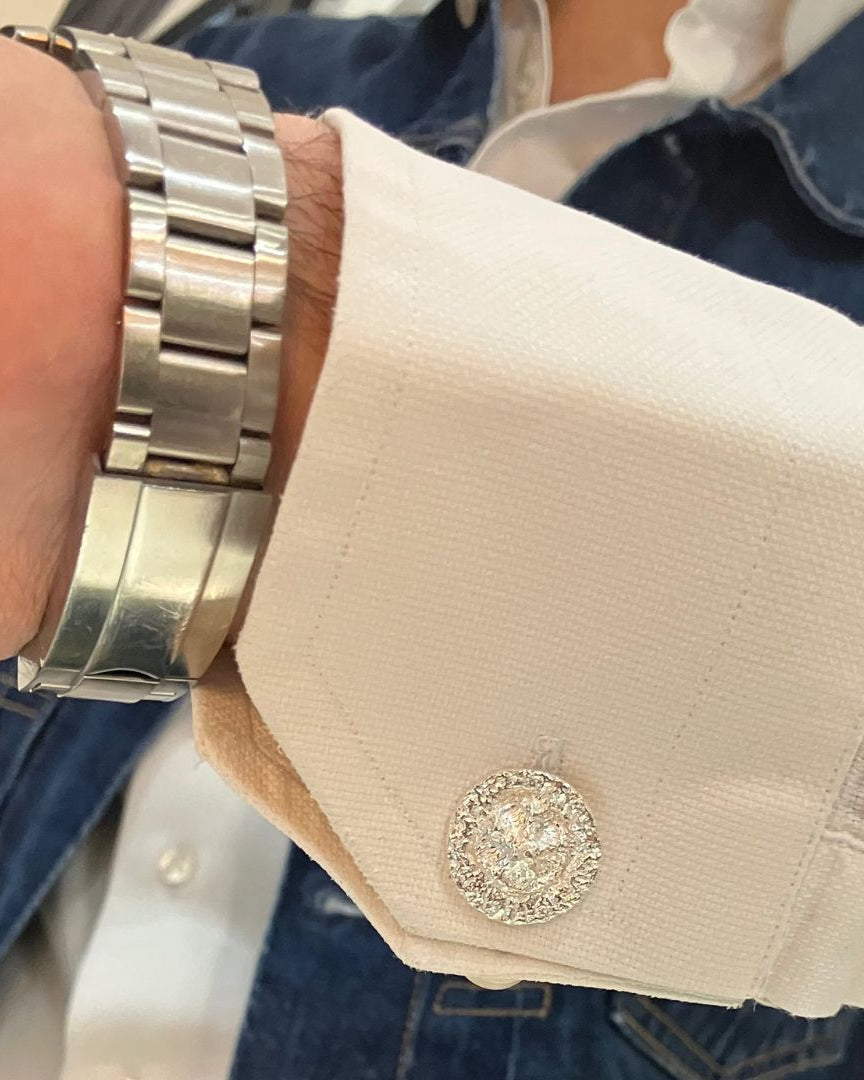 A close-up of a person's wrist wearing a silver metal link watch and a white dress shirt with a decorative round cufflink, layered over a blue denim jacket.