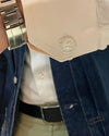 A close-up of a person's wrist wearing a silver metal link watch, with a cream-colored dress shirt cuff featuring a round, crystal-encrusted silver cufflink. A blue denim jacket and white collared shirt are visible in the background.