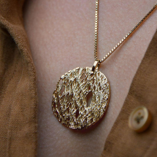 A gold textured pendant necklace worn on a person's chest over a brown button-up shirt.