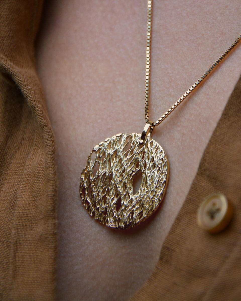 A gold textured pendant necklace worn on a person's chest over a brown button-up shirt.