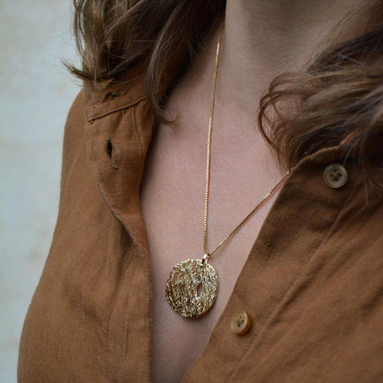 A close-up of a person wearing a textured gold pendant necklace on a delicate gold chain, paired with a brown button-down shirt.