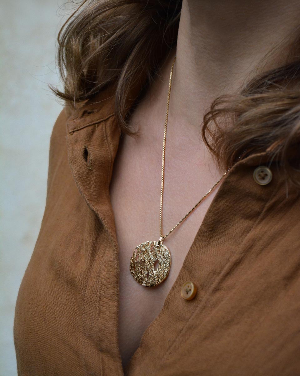 A close-up of a person wearing a textured gold pendant necklace on a delicate gold chain, paired with a brown button-down shirt.