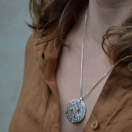 A close-up lifestyle shot of a person wearing a textured silver pendant necklace on a delicate silver chain, paired with a brown linen button-up shirt.
