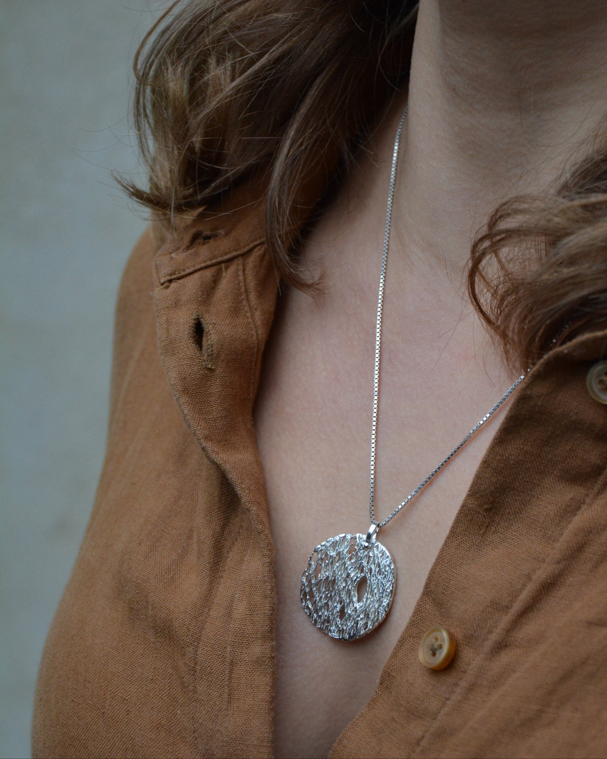 A close-up lifestyle shot of a person wearing a textured silver pendant necklace on a delicate silver chain, paired with a brown linen button-up shirt.