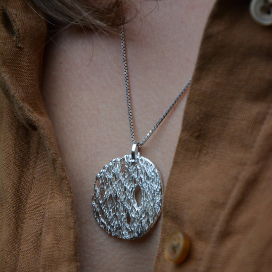 A silver textured oval pendant necklace worn on a person's neck over a brown linen button-up shirt.