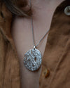 A silver textured oval pendant necklace worn on a person's neck over a brown linen button-up shirt.
