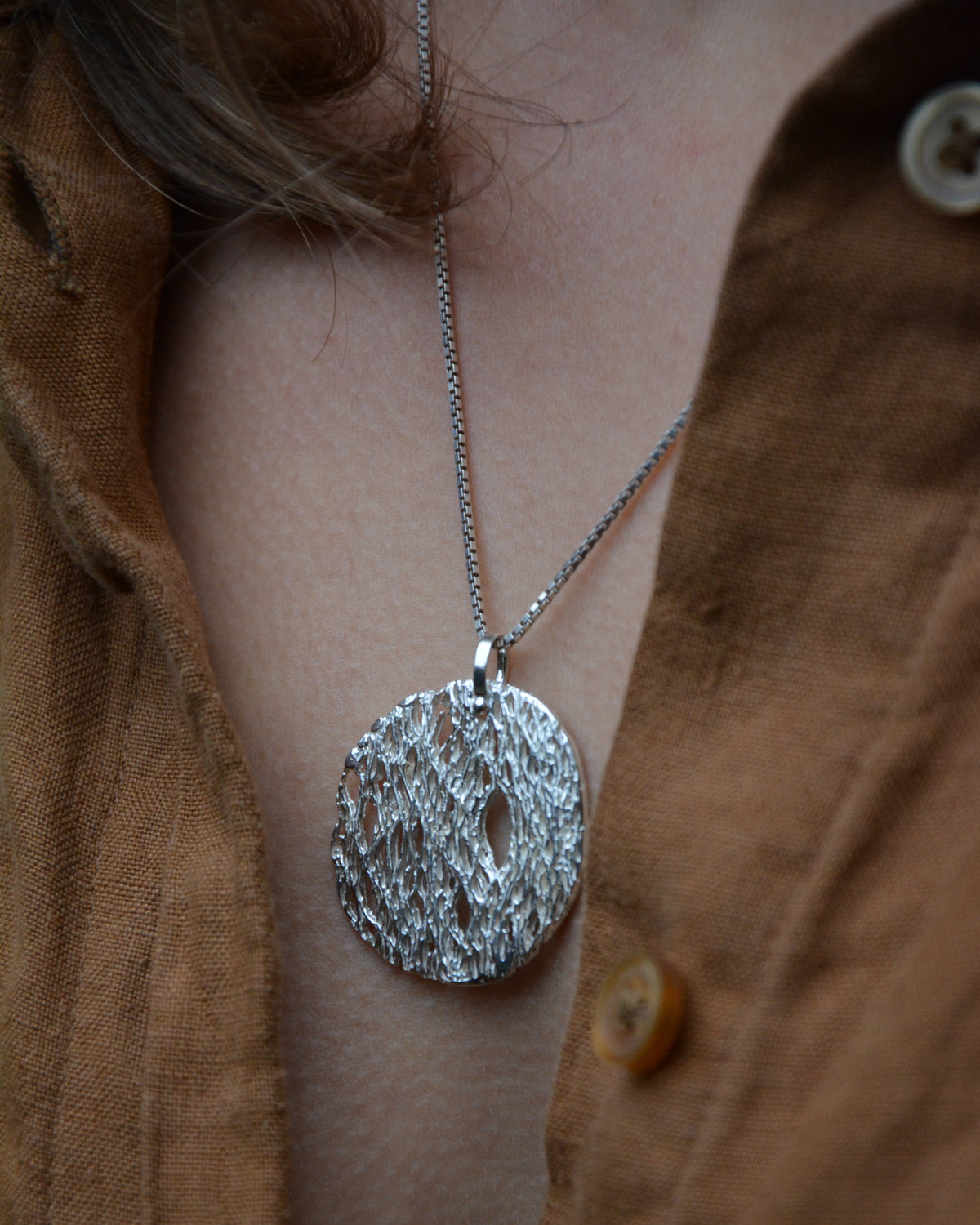 A silver textured oval pendant necklace worn on a person's neck over a brown linen button-up shirt.