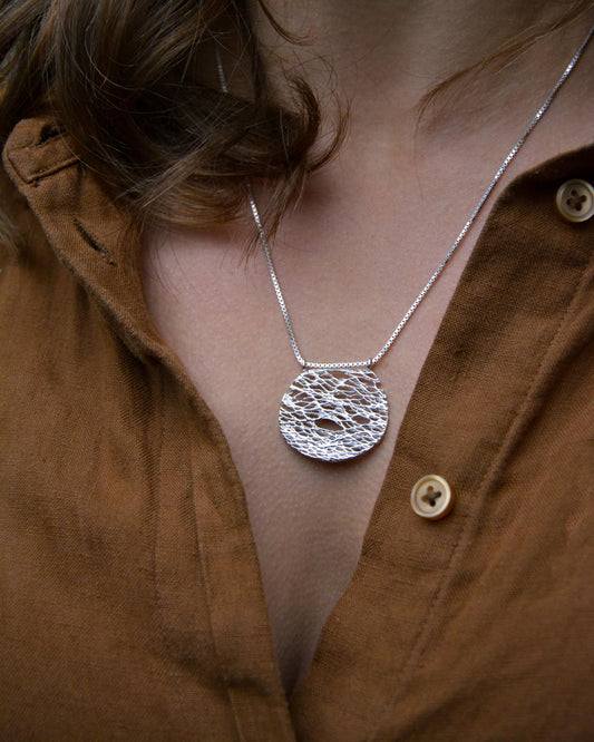 A close-up of a person wearing a silver pendant necklace with a textured, hammered circular disc design on a delicate chain, styled with a brown linen button-up shirt.