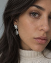 A close-up portrait of a woman wearing a silver drop earring with clear stones, a nose stud, and a cream cable-knit sweater.