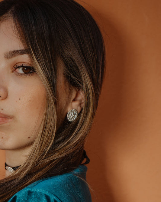 A close-up portrait of a woman with long brown hair wearing a textured teal top and a silver-toned geometric stud earring, set against a warm terracotta background.