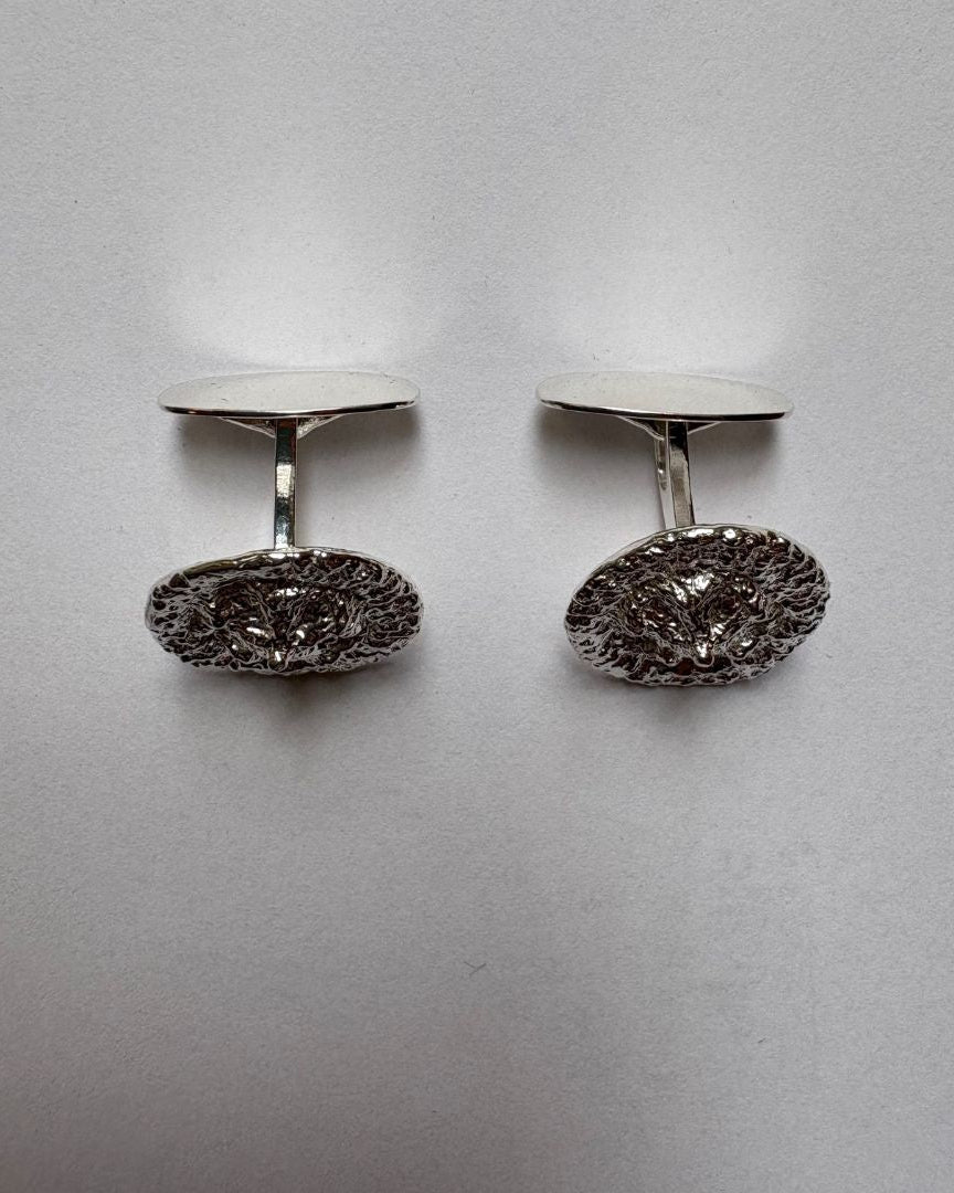 A pair of silver oval cufflinks with textured front, displayed on a neutral light gray background.