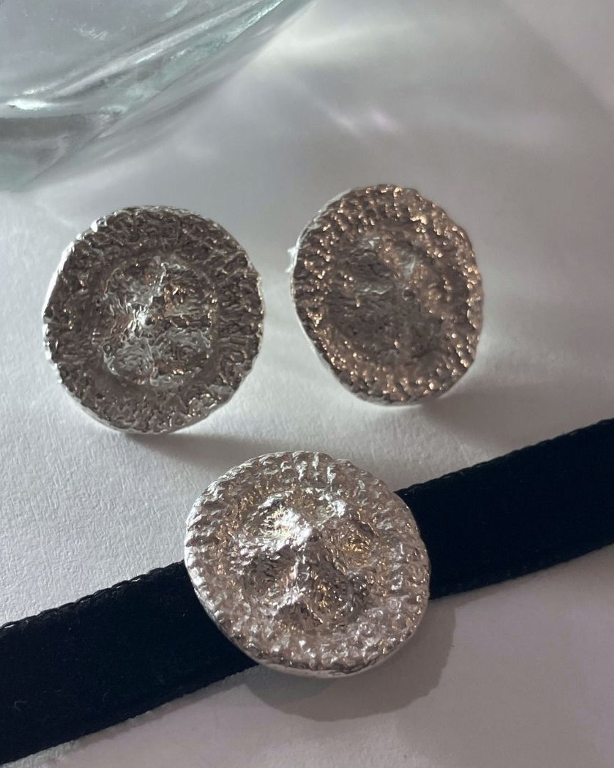 Three textured silver round stud earrings displayed on a white surface with a black ribbon and a glass bowl in the background.