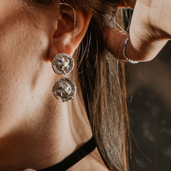 A close-up lifestyle shot of a woman wearing silver-toned circular drop earrings , a black choker with a matching round earring, and a silver ring on her tumb