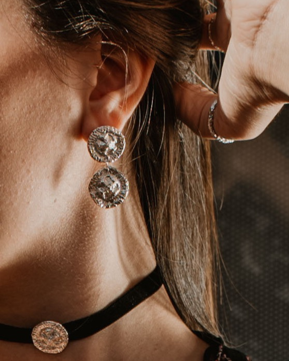 A close-up lifestyle shot of a woman wearing silver-toned circular drop earrings , a black choker with a matching round earring, and a silver ring on her tumb