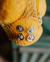 A close-up of a yellow lemon fruit, hanging in a yellow plastic mesh bag with two pendal silver circular studs