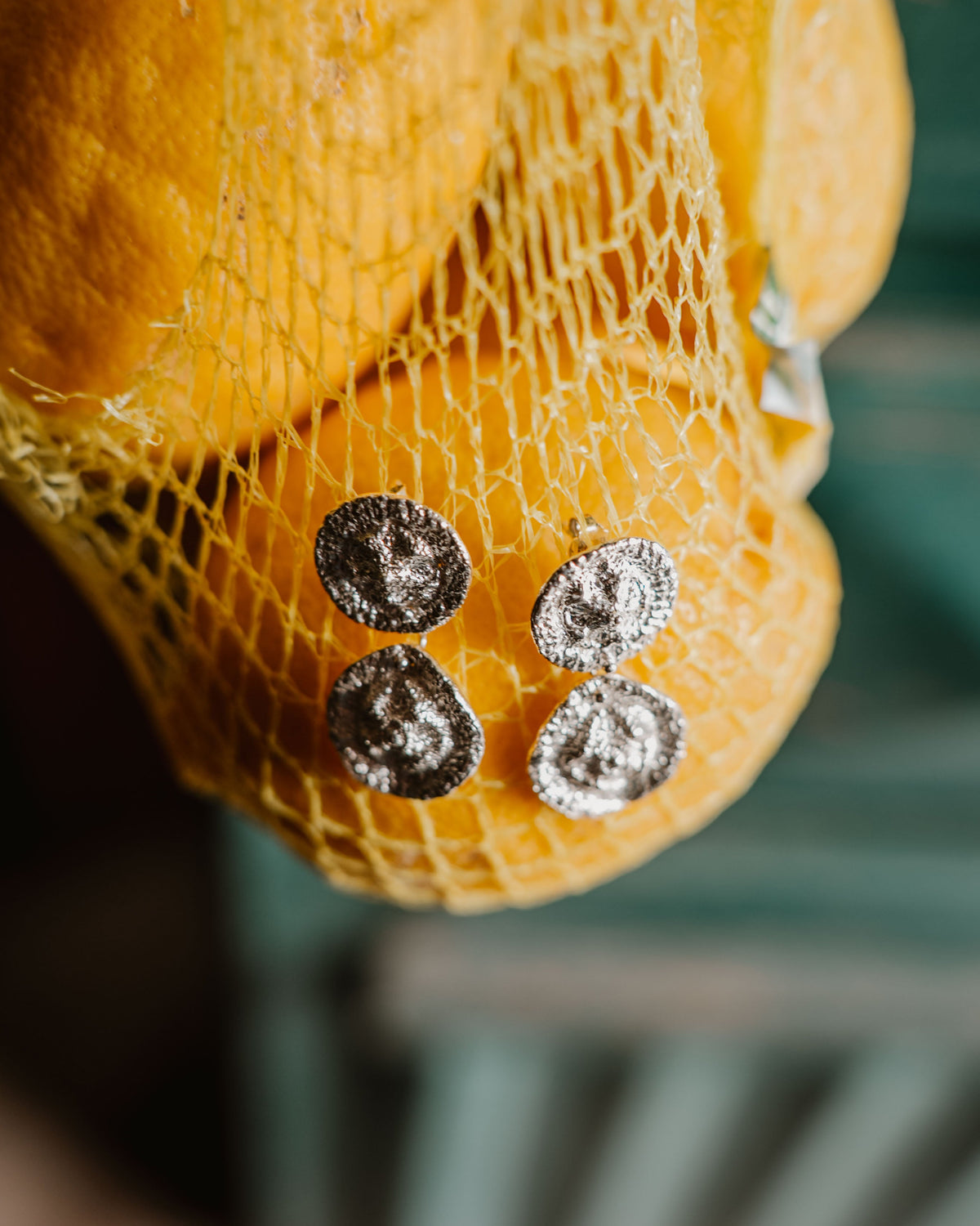 A close-up of a yellow lemon fruit, hanging in a yellow plastic mesh bag with two pendal silver circular studs