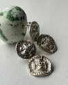 A close-up of textured silver coin-shaped earrings next to a green and white speckled stone on a light gray background.