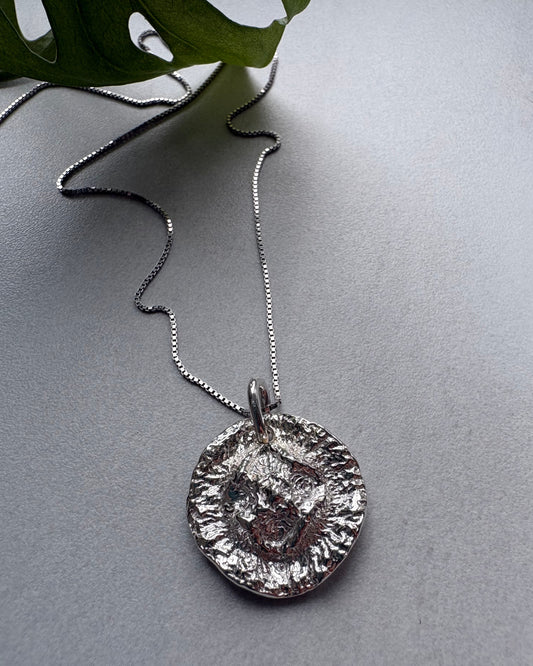 A textured silver pendant necklace with a circular, hammered finish on a matching chain, displayed on a gray surface with a green leaf in the corner.