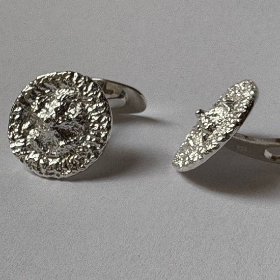 A pair of silver-toned cufflinks with textured, hammered circular faces, shown on a neutral gray background.