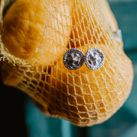 A close-up of a pair of silver round stud earrings resting on a yellow mesh bag containing oval lemon fruits, with a blurred teal background.