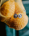 A close-up of a pair of silver round stud earrings resting on a yellow mesh bag containing oval lemon fruits, with a blurred teal background.