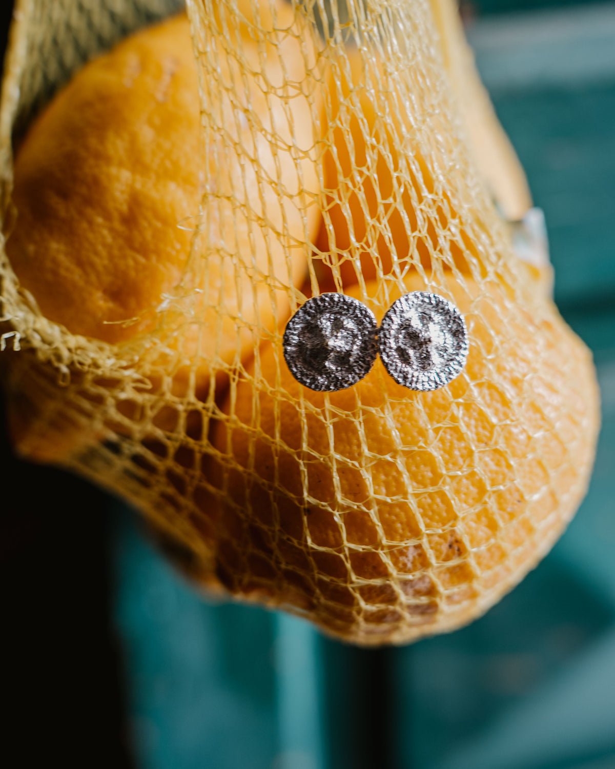 A close-up of a pair of silver round stud earrings resting on a yellow mesh bag containing oval lemon fruits, with a blurred teal background.