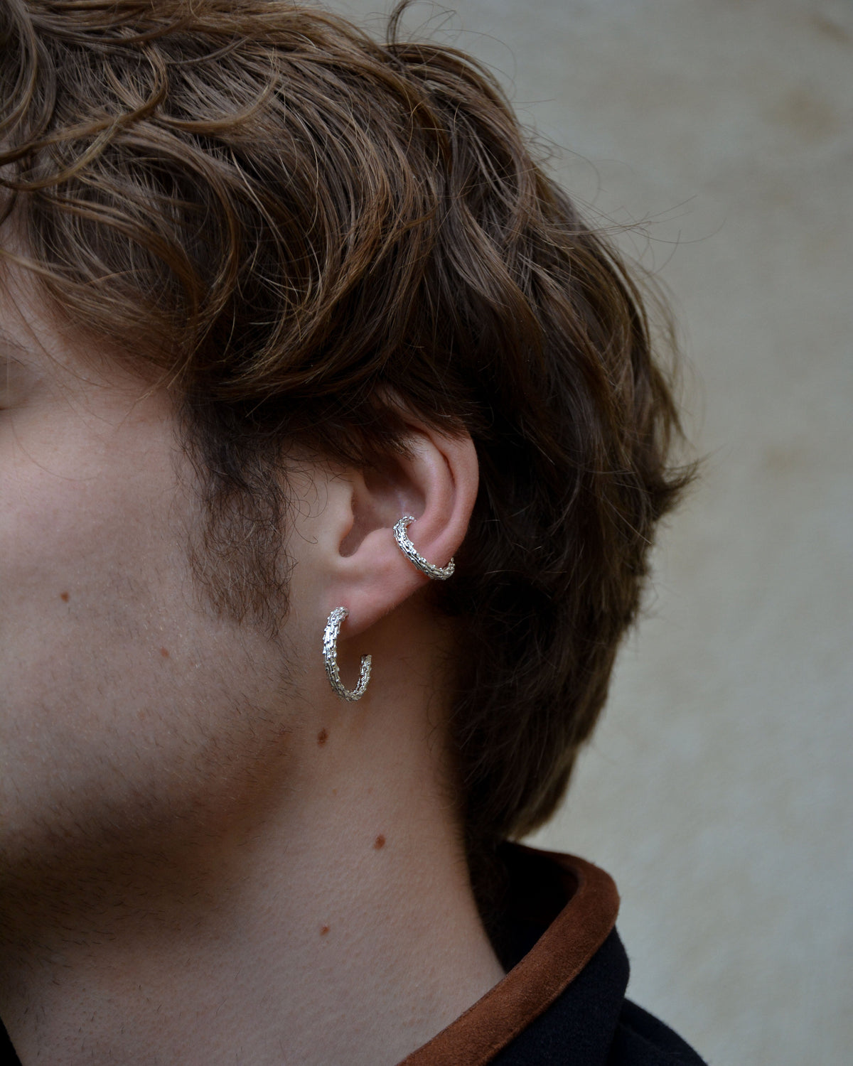 close up of model wearing a silver textured hoop and silver earcuff in the same style on a beige background