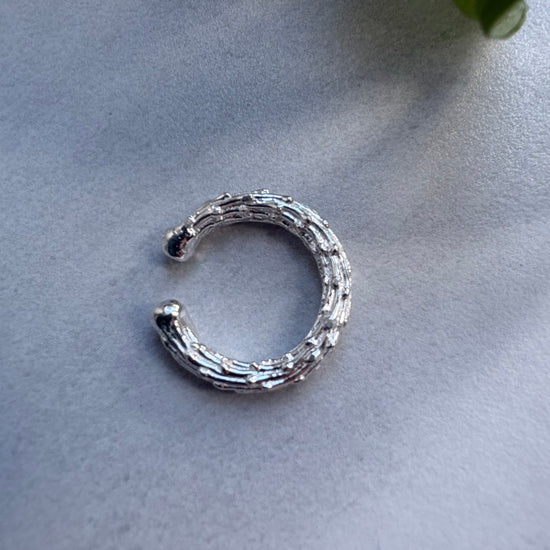 Silver earcuff on a light gray surface with a plant leaf in the corner.