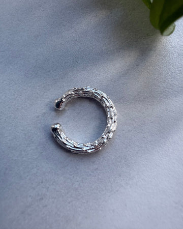 Silver earcuff on a light gray surface with a plant leaf in the corner.