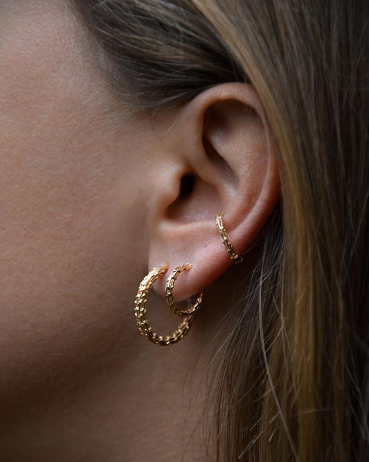 A close-up of a person's ear wearing textured gold hoop earrings and a matching gold ear cuff.
