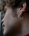 A close-up side profile of a person wearing two silver hoop earrings, one smaller textured hoop in the lobe and one larger textured hoop in the cartilage.