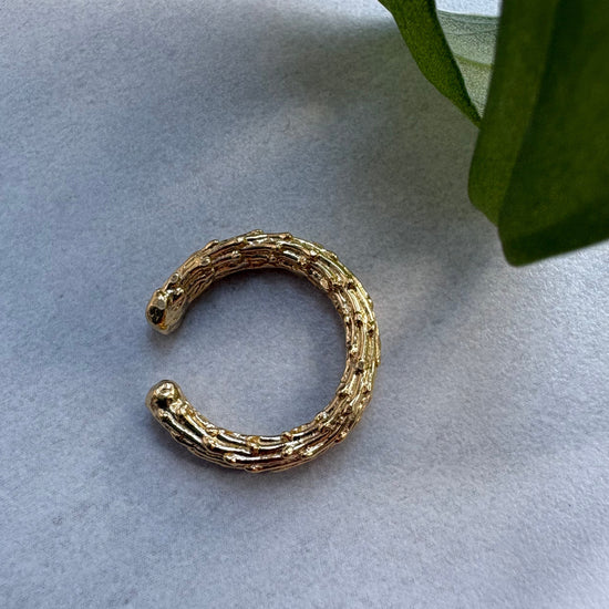 Gold organic ear cuff on a light gray surface with green leaves in the corner