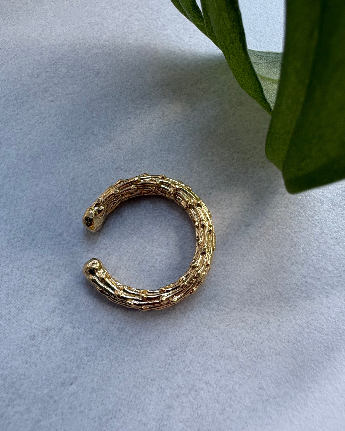 Gold organic ear cuff on a light gray surface with green leaves in the corner
