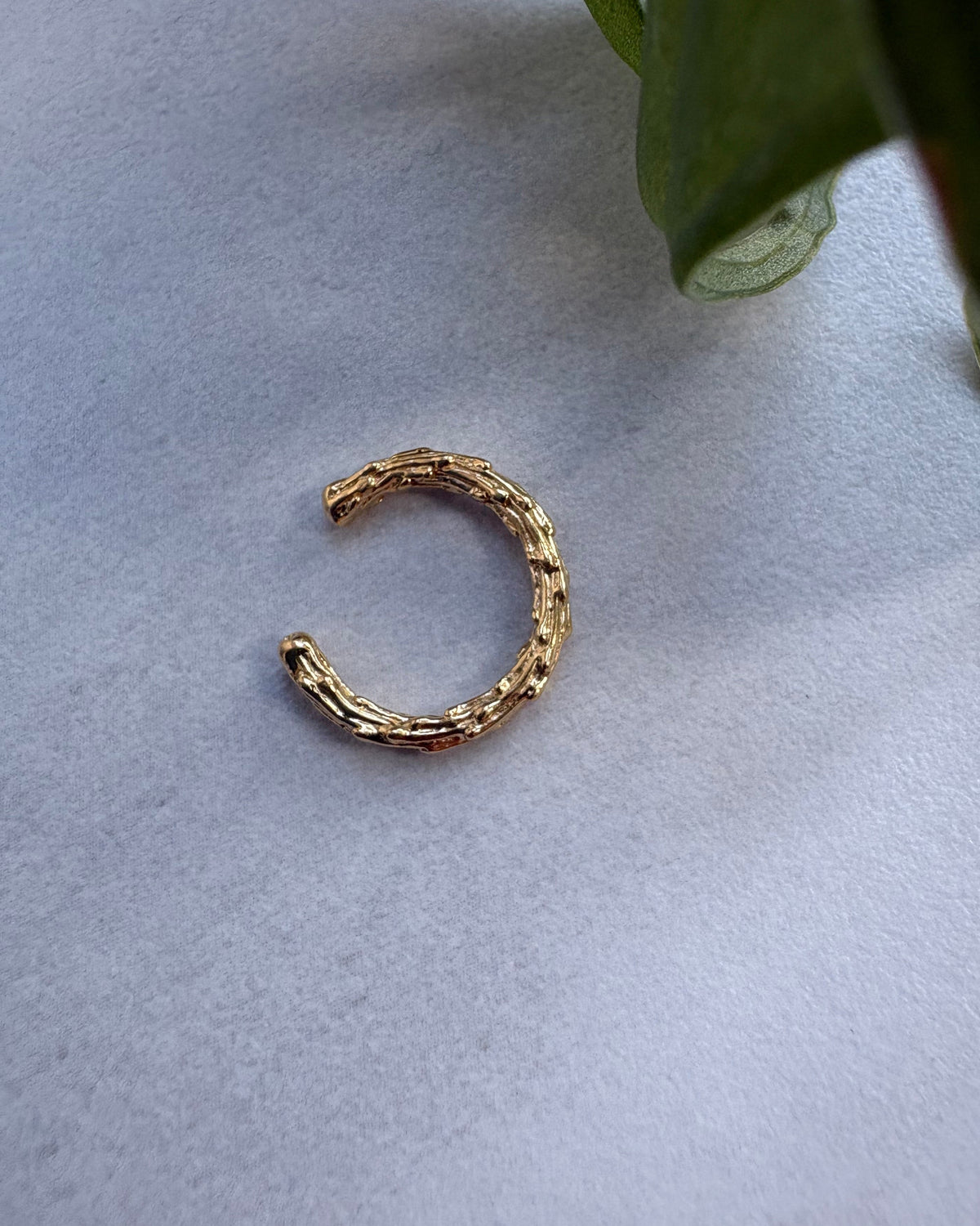 Goldplated earcuff on a light gray surface with a green leaf in the corner.