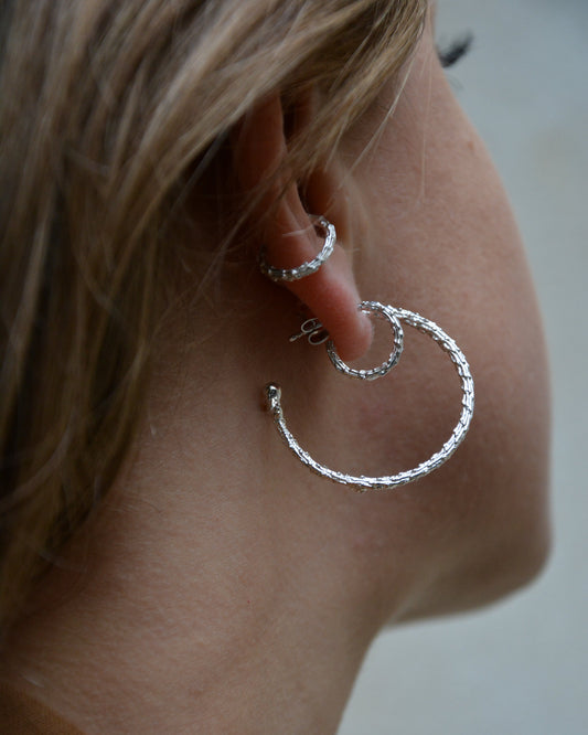 A close-up side profile of a person wearing textured silver hoop earrings, showing the ear, hair, and neck against a soft gray background.