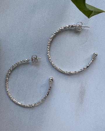 Silver hoop earrings on a light gray surface with a green leaf in the corner.