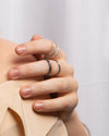 A close-up of a hand wearing four silver rings, including two delicate bands and two bigger textured ring, styled with a beige top against a soft white background.