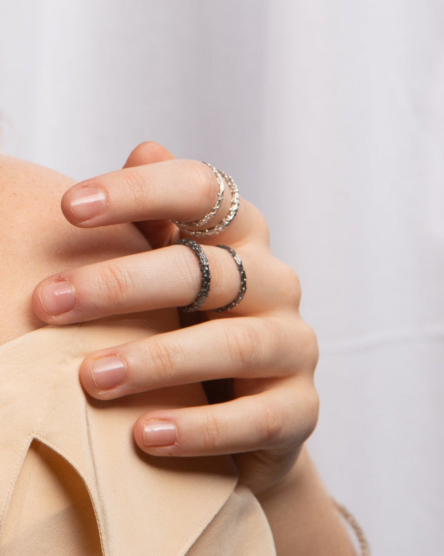 A close-up of a hand wearing four silver rings, including two delicate bands and two bigger textured ring, styled with a beige top against a soft white background.