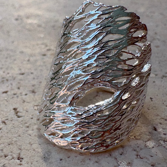 A textured, organic-shaped silver ring with a hollow, lace-like design, resting on a concrete surface outdoors.
