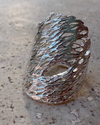 A textured, organic-shaped silver ring with a hollow, lace-like design, resting on a concrete surface outdoors.