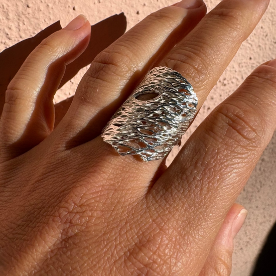 A close-up of a hand wearing a wide, textured silver ring with a hammered, organic surface pattern against a warm beige stucco wall.