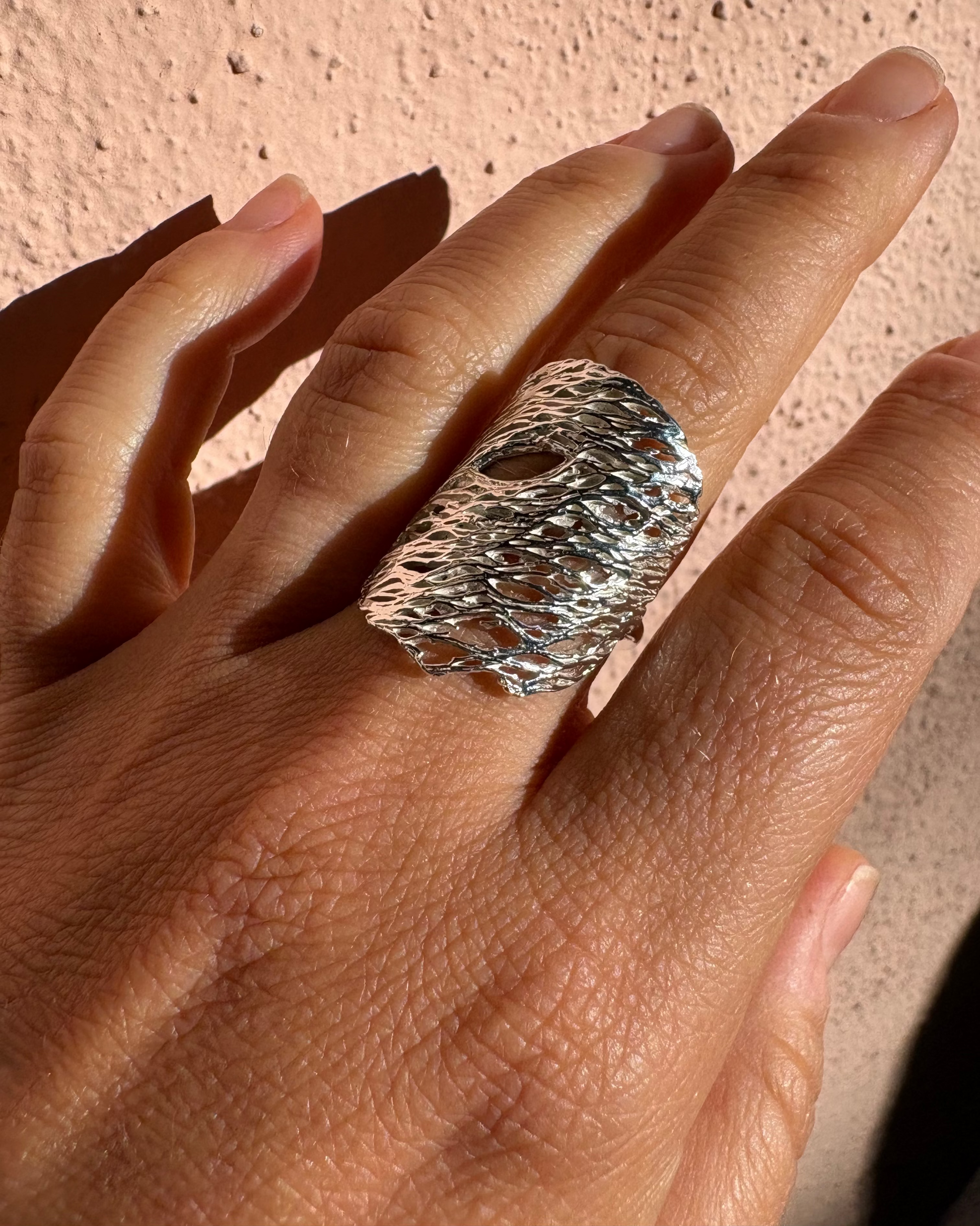 A close-up of a hand wearing a wide, textured silver ring with a hammered, organic surface pattern against a warm beige stucco wall.
