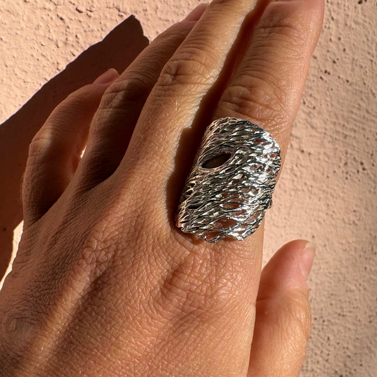 A close-up of a hand wearing a chunky, textured silver ring against a stucco wall background.