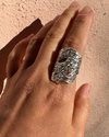 A close-up of a hand wearing a chunky, textured silver ring against a stucco wall background.