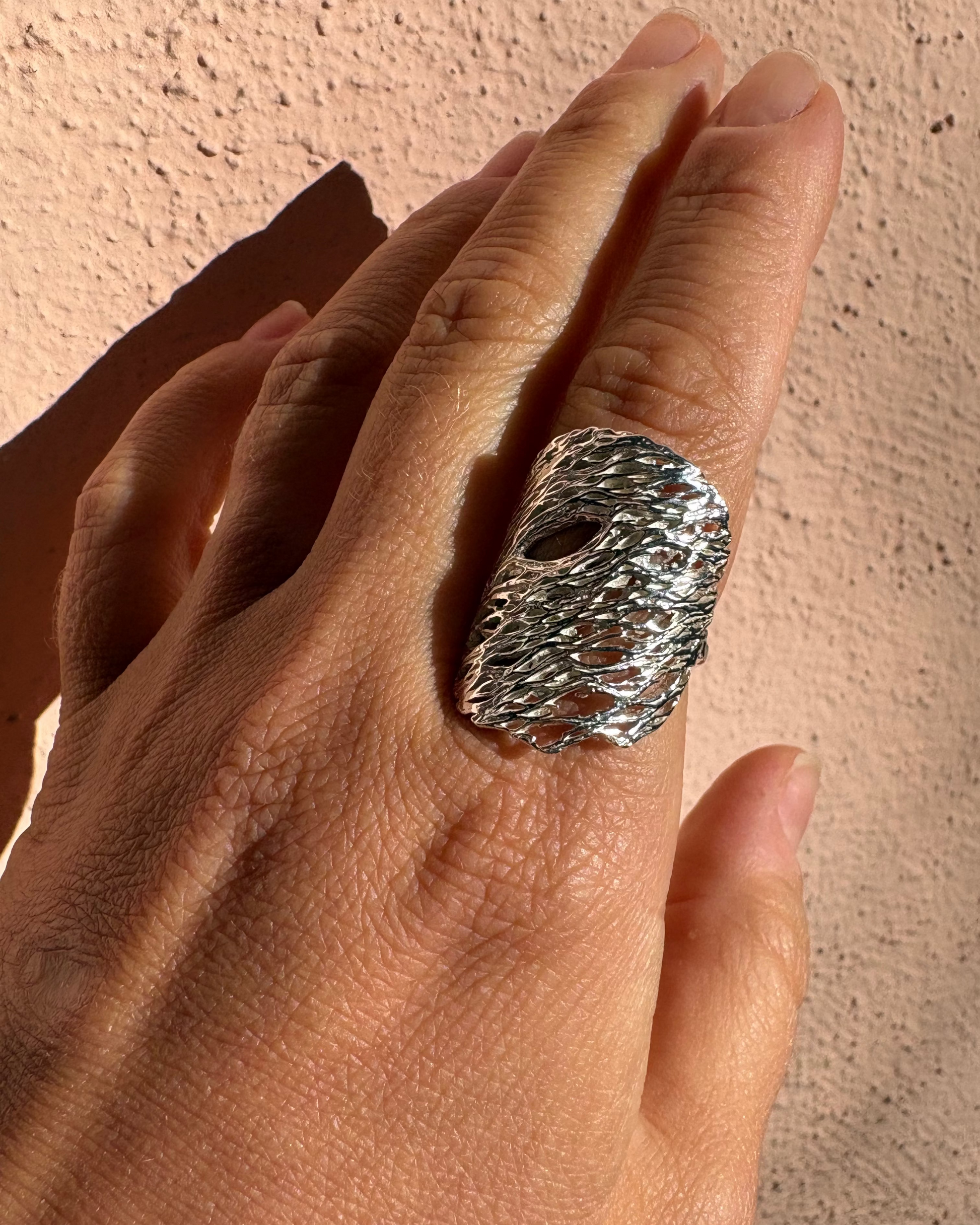 A close-up of a hand wearing a chunky, textured silver ring against a stucco wall background.