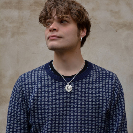 Person wearing a blue patterned sweater against a beige wall with a texture silver necklace