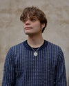 Person wearing a blue patterned sweater against a beige wall with a texture silver necklace