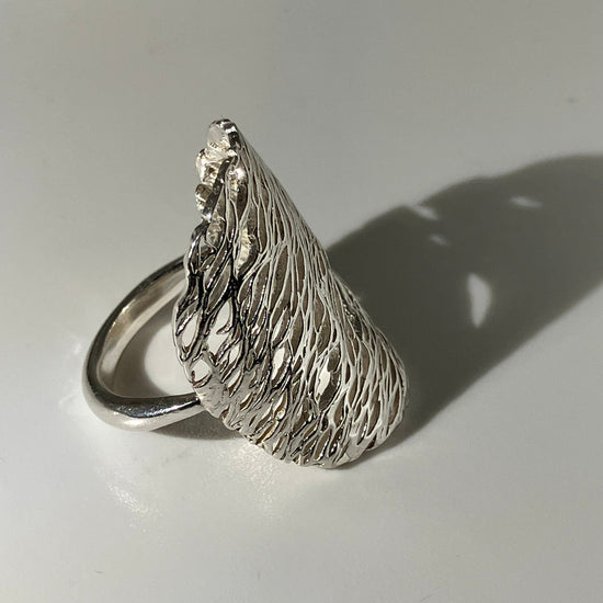 A textured sterling silver ring shaped like a stylized wing or leaf, photographed on a plain white surface with a sharp shadow.